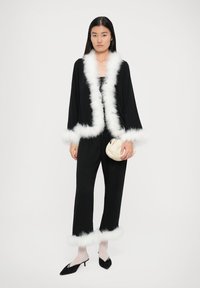 Outfit nero con rifiniture in pelliccia sintetica bianca su maniche e orli dei pantaloni. La modella tiene una pochette crema e indossa scarpe nere a punta e collant trasparenti.