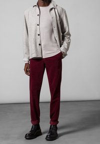 Šedý pletený kabát s černými knoflíky, béžová přiléhavá košile, bordó manšestrové kalhoty a černé kotníkové boty. Minimalistický design, bez vzorů.