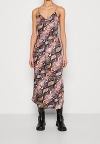 Femme portant une robe midi sans manches à motif serpent multicolore avec des fines bretelles et des bottes noires mi-mollet, se tenant devant un fond blanc.