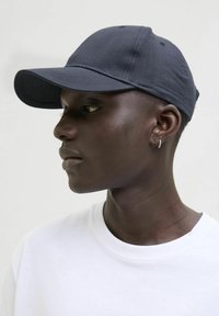 Casquette de baseball en tissu foncé avec une visière courbée, des coutures détaillées et une finition texturée, portée par une personne en chemise blanche.