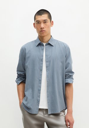 Homme portant une chemise boutonnée à rayures verticales bleues et blanches par-dessus un t-shirt blanc, manches retroussées et mains dans les poches, debout devant un fond blanc.