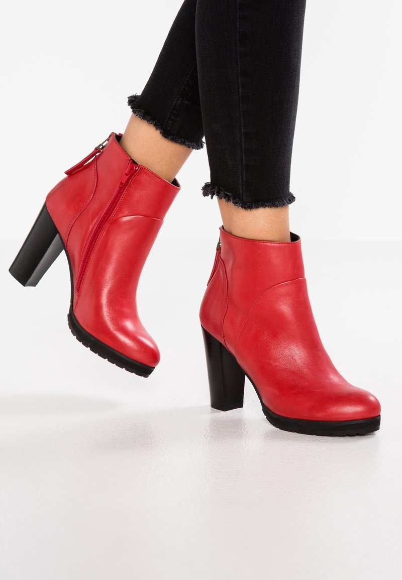 Bottes rouges en cuir à talons blocs, longueur cheville, portées avec un pantalon noir à ourlet effiloché.
