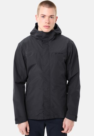 Junger Mann mit einer schwarzen, wasserdichten Kapuzenjacke mit Frontreißverschluss und Klettverschlüssen an den Handgelenken, stehend vor einem schlichten Hintergrund.