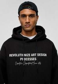 Zwarte hoodie met witte tekst "REVOLUTIONIZE ART DESIGN PROCESSES" en "Creative Department Innovates," inclusief een trekkoord in de capuchon.