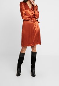 Satiiniwrap-mekko poltettuoranssissa, jossa on pitkät hihat ja napilliset hihansuut; yhdistettynä polveen saakka ulottuviin mustiin mokkanahkabootseihin, joissa on nahka-akseli.