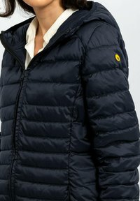 Giacca a vento imbottita blu navy con cappuccio, caratterizzata da cuciture orizzontali, cerniera frontale e un piccolo logo giallo rotondo sulla manica sinistra.