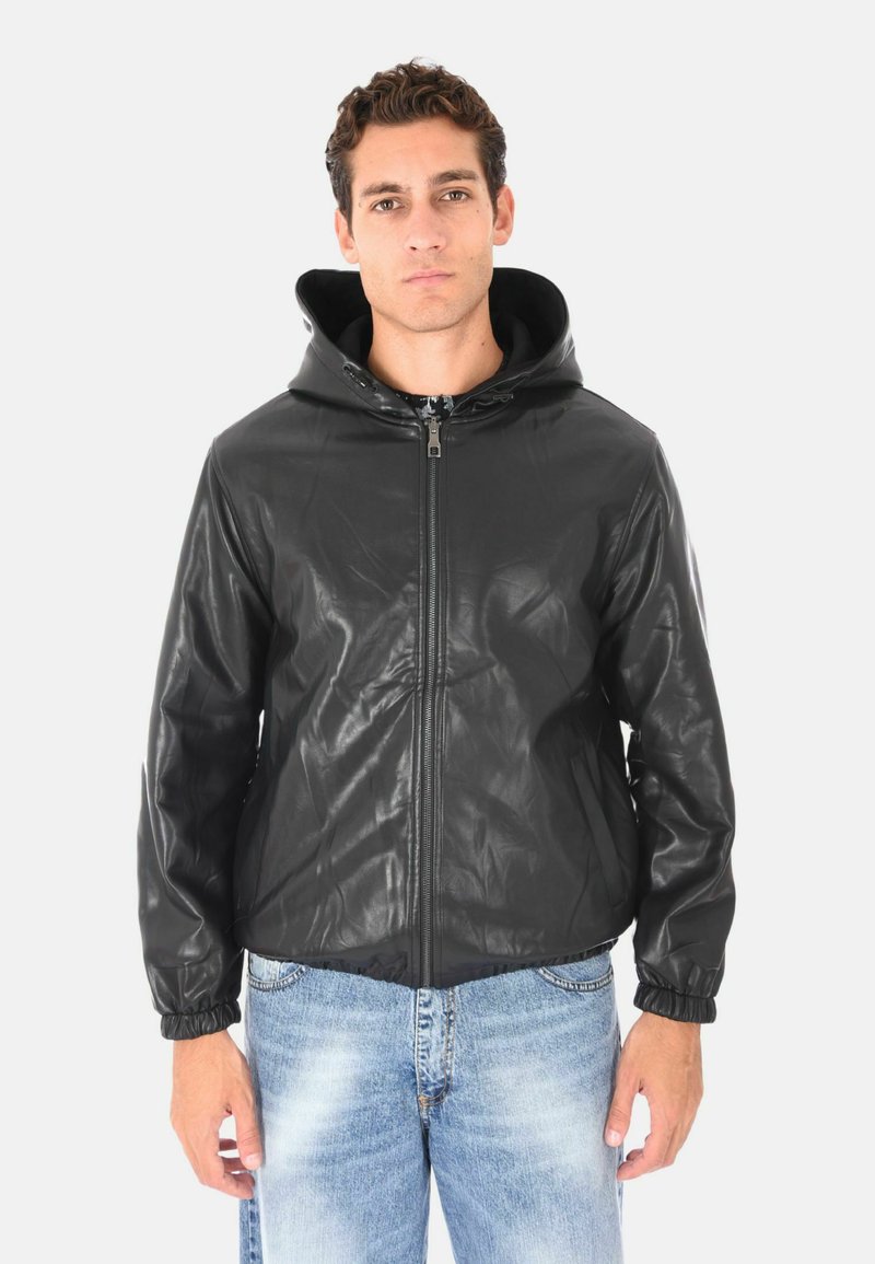 Uomo che indossa una giacca di pelle nera con zip e cappuccio e jeans chiari sbiaditi blu, in piedi contro uno sfondo bianco semplice.