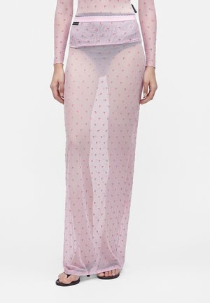 Modèle portant une jupe longue rose transparente et un haut avec de petits motifs rouges en forme de croissant, associé à des sandales plates noires, debout devant un fond uni.