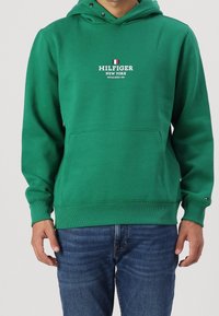 Grüne Baumwollkapuzenjacke mit einer Fronttasche, Logo-Text "HILFIGER NEW YORK" und gerippten Bündchen und Saum. Kombiniert mit blauen Jeans.