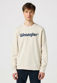 Felpa beige chiaro con scollo rotondo e maniche lunghe. Presenta il logo "Wrangler" in blu navy in evidenza sulla parte anteriore. Comodo materiale in cotone.