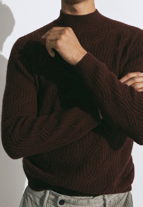 Personne portant un pull en maille torsadée marron foncé, les bras croisés et une main près du menton, assorti à un pantalon clair.