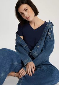 Granatowa koszulka z dekoltem w serek pod niebieską dżinsową kurtką. Model siedzi w stylowych dżinsowych spodniach o szerokich nogawkach, odsłaniając postrzępione krawędzie i luźny krój.