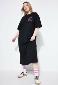 Czarna, oversize sukienka T-shirt z krótkimi rękawami, z różową grafiką "Live Love", bocznymi rozporkami, noszona z skarpetami w różowe paski i trampkami.
