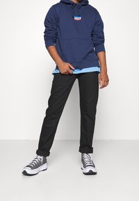 Felpa con cappuccio blu navy con logo Levi's rosso e blu, indossata sopra una camicia azzurro chiaro, abbinata a pantaloni neri e sneaker grigie con dettagli bianchi.