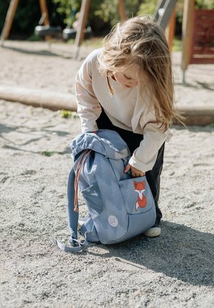 Zaino azzurro chiaro con design di volpe, chiusura con cordoncino, tasche spaziose e spallacci regolabili, posato su un terreno sabbioso.