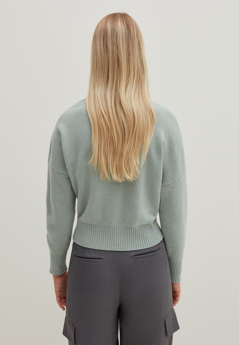 Hellgrüner Strickpullover mit geripptem Saum und Manschetten, der eine lockere Passform hat. Über grauen Hosen getragen, mit Haaren, die den Rücken hinabfallen.