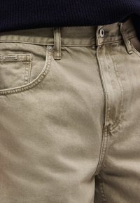 Κοντινό πλάνο μπεζ βαμβακερών παντελονιών με κουμπί και φερμουάρ, ορατή μπροστινή τσέπη, θηλιές για ζώνη και μια μικρή ετικέτα «LINDBERGH» στην άκρη της τσέπης.