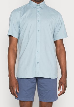 Mann trägt ein hellblaues, kurzärmeliges Hemd mit Knöpfen und dunkelblaue Shorts vor neutralem Hintergrund.