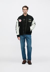 Νέος άνδρας που φοράει μαύρο και άσπρο τζάκετ με φερμουάρ και μπαλώματα, μπλε τζιν και καφέ παπούτσια, στέκεται μπροστά σε απλό φωτεινό φόντο.