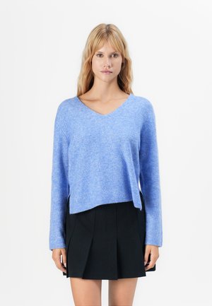 Vrouw in een lichtblauwe V-hals trui en een zwarte geplooide rok, staande tegen een witte achtergrond, recht vooruit kijkend.