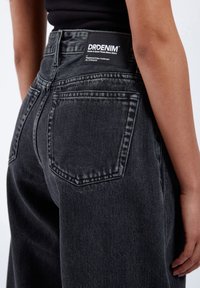 Person som bär hög midja svarta denimjeans med en DRDENIM-etikett på midjebandet och en synlig bakficka på höger sida.