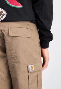 Gros plan sur une personne portant un pantalon cargo Carhartt beige avec poches à rabat et une partie d'une chemise noire à imprimé de fruits.