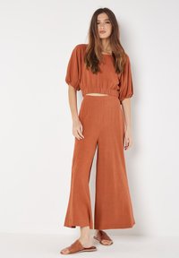 Roestkleurige twee-delige outfit bestaande uit een cropped top met geplooide mouwen en brede broekspijpen. Zachte, lichte stof met een gladde textuur.