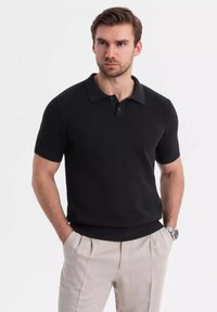 Černé polo tričko s krátkým rukávem a texturovaným pleteným vzorem, s klasickým límcem a zapínáním na dva knoflíky, v kombinaci s světle zbarvenými kalhotami.