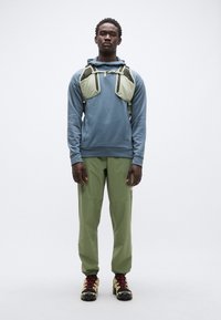 Αντιανεμικό σε μπλε χρώμα με υφασμάτινη υφή, σε συνδυασμό με ανοιχτό πράσινο παντελόνι και μαύρα παπούτσια με κόκκινα κορδόνια. Στο μπροστινό μέρος υπάρχει ένα ιμάντας με αντίθετο χρώμα.