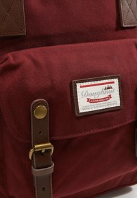 Bordowy plecak z płótna z brązowymi skórzanymi akcentami, mosiężnymi elementami i haftowaną etykietą. Posiada teksturowaną powierzchnię i przednią kieszeń.