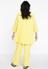 Geel driedelig ensemble met loszittende top en broek met wijd uitlopende pijpen, gemaakt van een getextureerde stof. Het ontwerp heeft driekwart mouwen en een ontspannen silhouet.