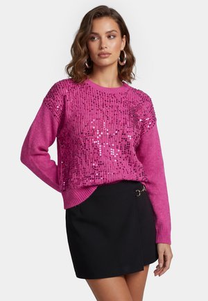 Kvinde med skulderlangt hår iført en magenta paliettrøje og sort nederdel, poserer med den ene hånd på hoften mod en hvid baggrund.