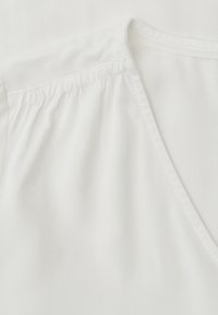 Vêtement blanc avec couture froncée à l'épaule et encolure en V, montrant des détails de couture fins sur un tissu lisse.