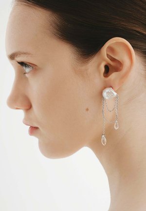 Perfil lateral de mujer con pendientes de cadena plateada con tachuelas de perla blanca y gotas similares a cristales sobre un fondo blanco liso.