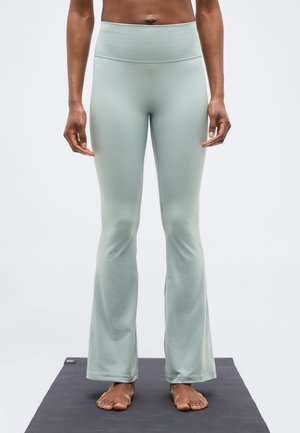 Osoba stojaca naboso na jogovej podložke, oblečená v svetlosivých rozšírených legínach a s odhaleným bruškom na jednofarebnom pozadí.