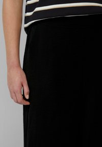 Zwarte stoffen broek met een gestructureerd oppervlak, gecombineerd met een zwart-wit gestreept bovenstuk, met een zichtbaar hand rustend nabij de taille.