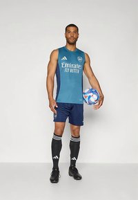 Zils bezroku krekls ar "Emirates" logotipu, tumši zili šorti, melnas ceļa zeķes ar baltām svītrām un melni futbola apavi, turot futbola bumbu.