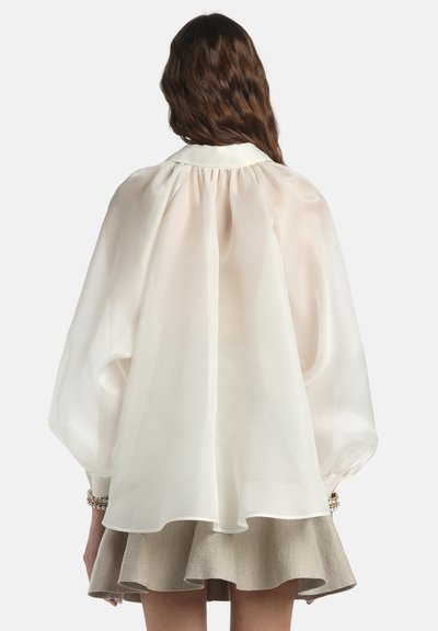 Sieviete ar viļņainiem brūniem matiem valkā caurspīdīgu, brīvi piegulošu baltu blūzi ar pūstām piedurknēm un bēšu izplešamu svārku, skatoties no muguras.