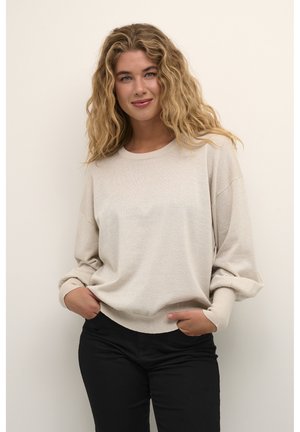 Pull à manches longues beige avec une coupe décontractée, un col rond et des poignets côtelés. Le matériau semble doux avec un léger éclat. Associé avec un pantalon noir.