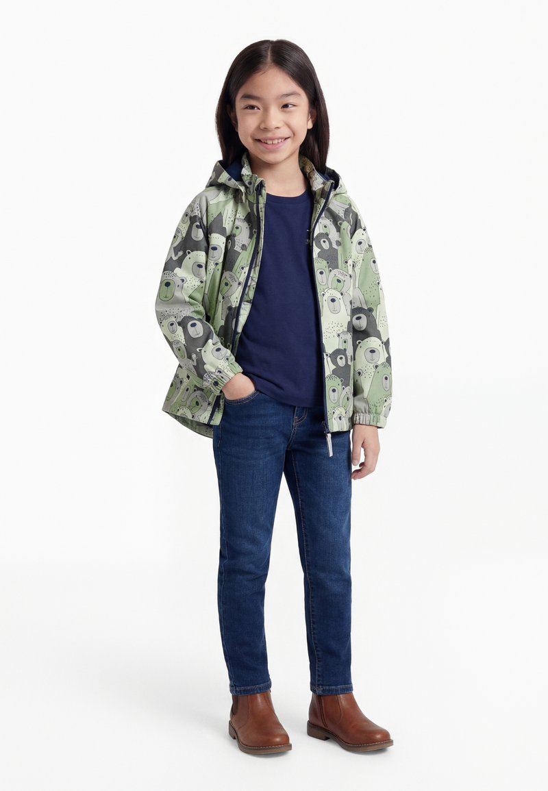 Fille souriante portant une veste verte à motifs d'ours, un chemisier bleu marine, un jean bleu et des bottes marron, debout avec une main dans la poche.