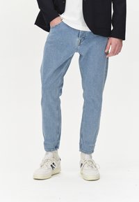 Jeans de mezclilla azul claro con un corte relajado, cinco bolsillos y un sutil detalle de costura, combinados con zapatillas blancas y un blazer negro.