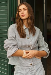 Jeune femme aux cheveux bruns lâchés appuyée contre une fenêtre à volets verts, portant une chemise boutonnée rayée bleu et blanc et un pantalon assorti.