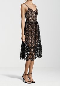 Robe en dentelle noire avec un décolleté en V, des bretelles fines et un ourlet à hauteur du genou. Elle présente des motifs floraux complexes et un corsage ajusté.