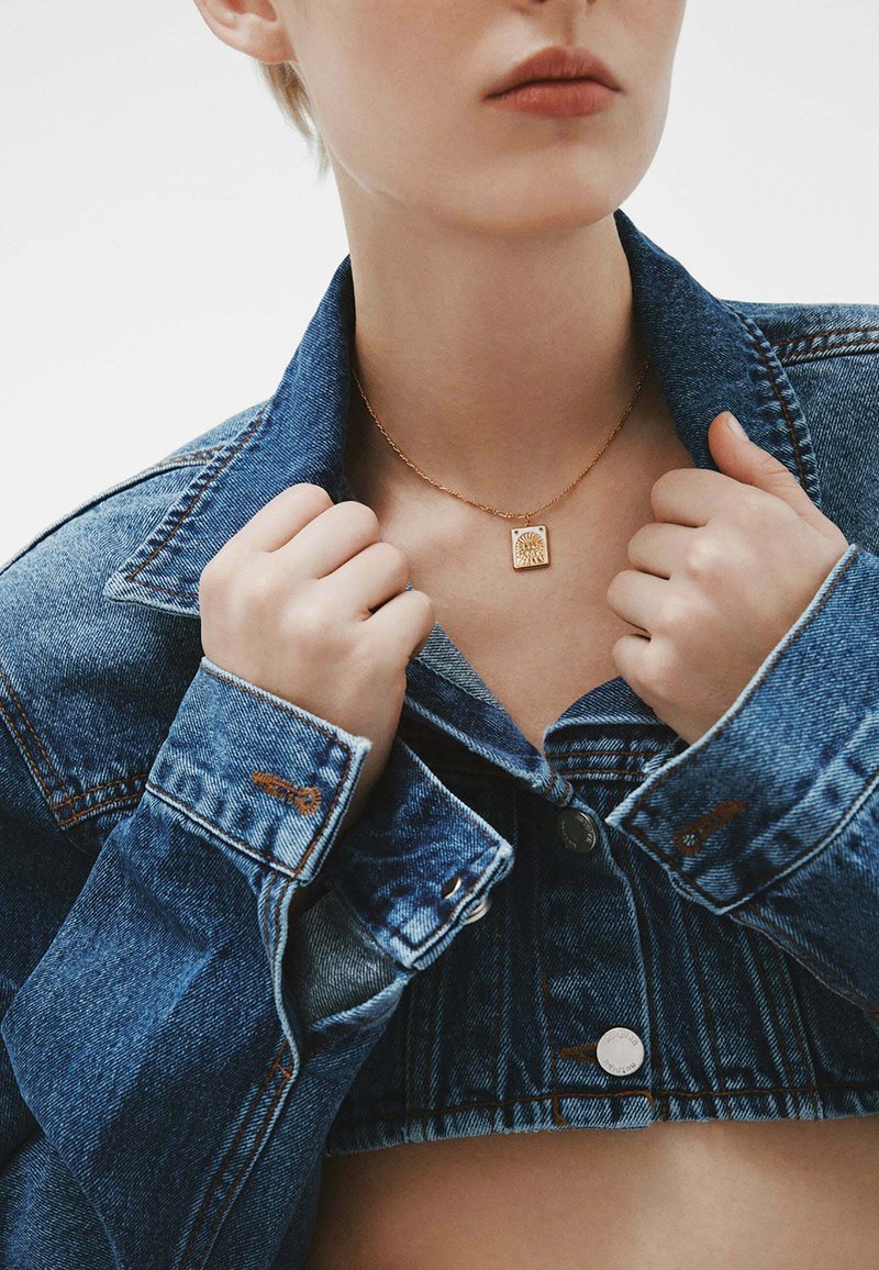 Collar de oro con un diseño cuadrado que presenta un patrón texturizado, llevado sobre una chaqueta de mezclilla con un cuello clásico.