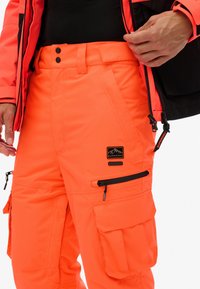Heldere oranje cargo-broek met meerdere zakken en zwarte ritsdetails, gedragen met een bijpassende oranje en zwarte jas.