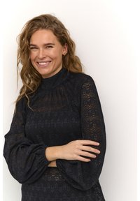 Black lace dress with high neck and voluminous sleeves. Textured pattern with sheer accents. Model poses with arms crossed against a plain background.