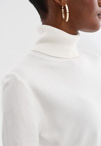 Zbliżenie na osobę w białym prążkowanym swetrze z golfem i dużych złotych kolczykach w kształcie koła, widoczną z boku na jednolitym tle.