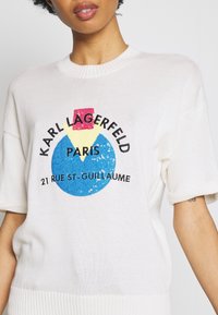 Bílý pletený svetr se zajímavým grafickým logem skládajícím se z modrého kruhu, růžového čtverce a žlutého trojúhelníku. Žebrovaný límec a manžety.