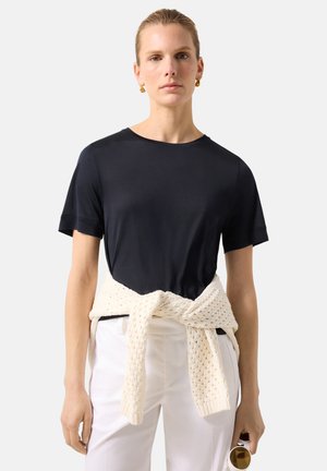 Kobieta z upiętymi włosami, ubrana w czarną koszulkę, białe spodnie, kremowy sweter z dzianiny zawiązany w talii i trzymająca okrągłe okulary przeciwsłoneczne.