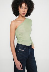 Top verde claro de un solo hombro con detalles fruncidos, combinado con vaqueros de mezclilla azul oscuro que presentan un bolsillo parche y tela texturizada.